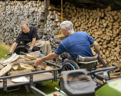 Two men in wheelchairs cut fire wood.