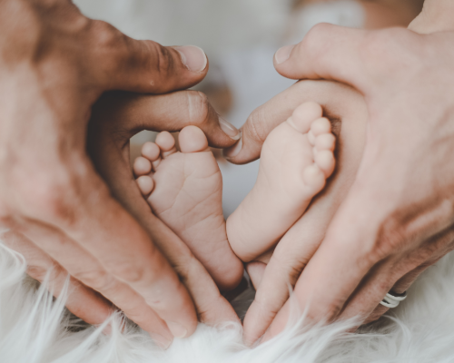 Two sets of hands making the shape of a heart around a baby's feet.