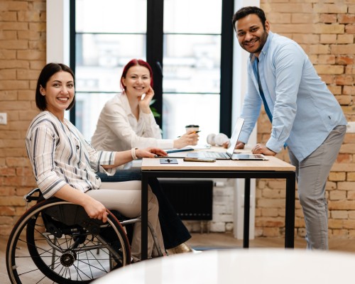 Three people sitting around a work desk smiling at the camera. A female worker is using a wheelchair.