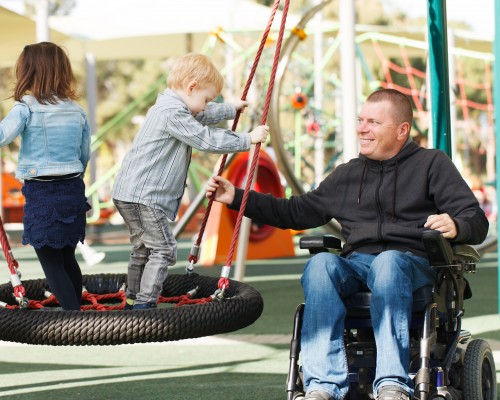 Dad using wheelchair playing with two young children on a swing