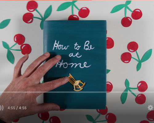 The background is cherries on a tablecloth. Sitting on the table cloth is a book entitled How to Be Alone. On the book is a tiny rocking  chair. A human hand is across the image reaching over to rock the tiny chair on the book