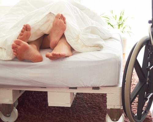 Two people in bed with feet wrapped together - a wheelchair is at the side of the bed